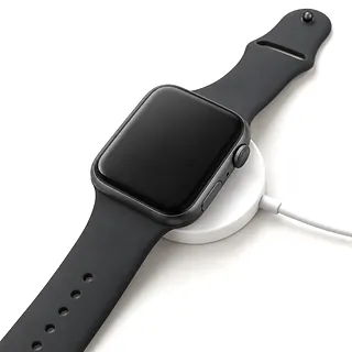 Apple Watch in dark gray with a black band, face up and off-center on a white charging puck with a dark screen